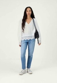 Vit blus med V-ringning och spetsdetaljer, ihop med ljusblå jeans. Modellen bär vita sneakers och har en svart väska.
