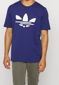 Námořnická modrá bavlněná tričko s velkým bílým logem Adidas se třemi pruhy a listovými akcenty. Krátké rukávy, design s kulatým výstřihem.