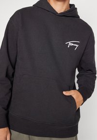 Felpa nera in cotone con tasca a marsupio, polsini e orlo a costine. Presenta un logo bianco "Tommy" sul petto. Texture morbida.