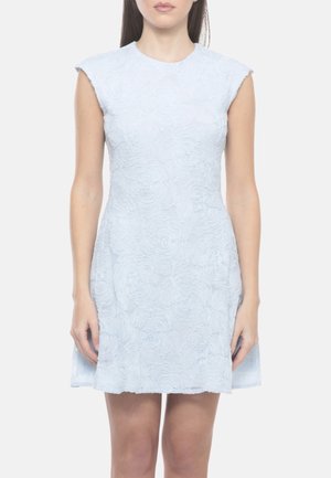 Donna che indossa un vestito azzurro chiaro senza maniche con motivi floreali strutturati, in piedi davanti a uno sfondo bianco uniforme.