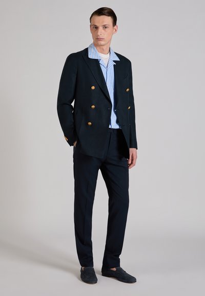 Junger Mann steht, trägt einen schwarzen zweireihigen Blazer mit goldenen Knöpfen, blau kariertes Hemd, schwarze Hose und schwarze Loafers.