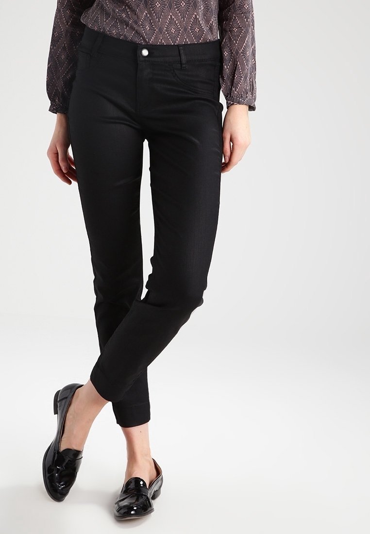 Femme porte un pantalon noir slim avec des mocassins en cuir verni noir et un haut à manches longues à motifs, debout jambes croisées.