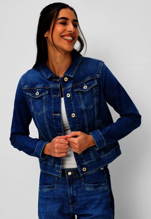 Femme souriante, ajustant un bouton sur une veste en jean bleu foncé, portant un jean assorti et une chemise blanche sur un fond clair uni.