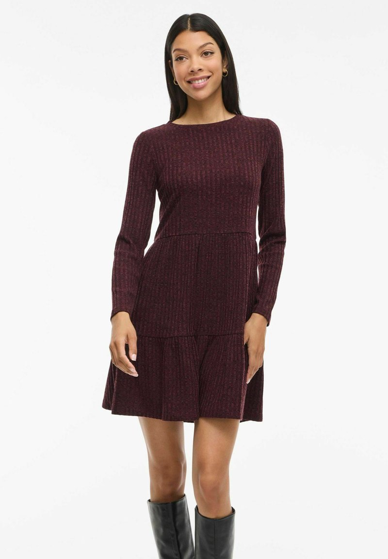Robe tricotée bordeaux avec des manches longues, texture côtelée, encolure ronde et détail de jupe à volants. Portée avec des bottes noires jusqu'aux genoux.