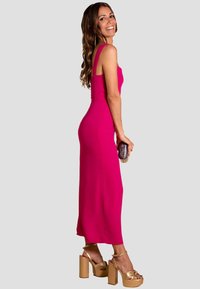 Vestido ajustado fucsia con tirantes finos, de longitud midi, con textura suave. Combinado con tacones de plataforma dorados y sosteniendo un clutch.