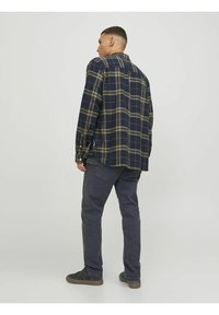Camisa de cuadros en tonos azul marino y oliva, de manga larga y botones, combinada con pantalones gris oscuro y zapatos negros, vista desde la parte posterior.