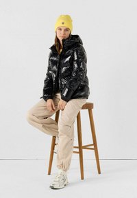 Giacca a vento nera lucida con cappuccio, pantaloni leggeri beige e sneakers argentati; modello seduto su uno sgabello di legno, berretto giallo.