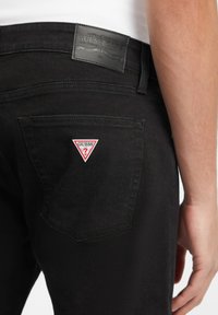 Svarte denimjeans har en baklomme med et rød og hvit trekantet "GUESS?" logo-lapp. Livsnormen har en teksturert svart lærmerke.
