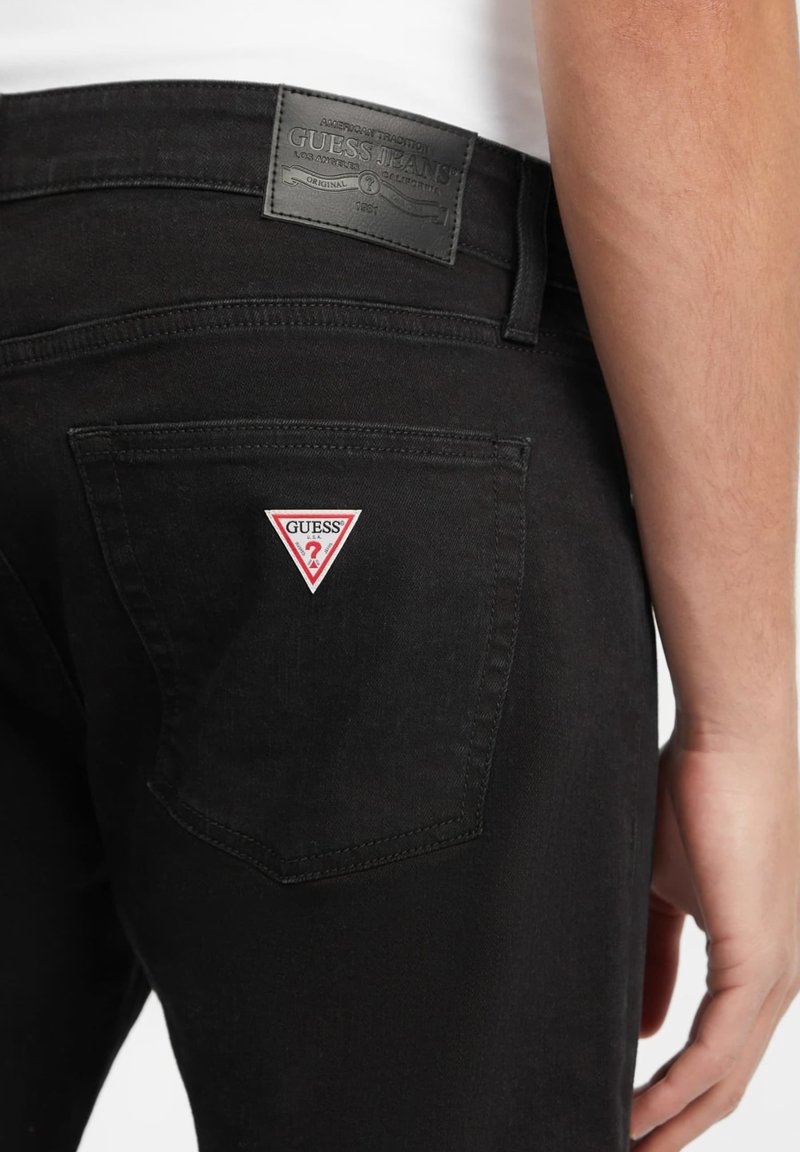Les jeans en denim noir présentent une poche arrière avec un patch en forme de triangle rouge et blanc portant le logo "GUESS?". La ceinture est ornée d'une étiquette en cuir noir texturé.