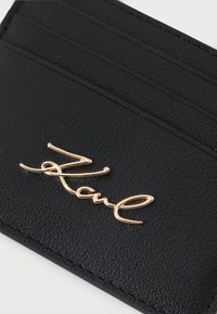 Melnas ādas karšu turētājs ar vairākiem karšu slotiem un zeltītajām firmas logo iegravēts priekšpusē. Gluda virsma ar izšūtu malām.