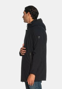 Giacca nera con cappuccio e chiusura con zip, caratterizzata da una texture liscia e tasche laterali, indossata da una persona di lato.