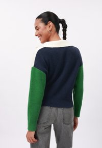 Knit sweater with a high collar, featuring navy, cream, and green blocks. Textured fabric. Paired with gray high-waisted jeans.