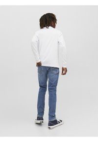 White long-sleeve shirt, blue jeans, and navy shoes. Features a relaxed fit with minimal design. Back view shows shirt without prints.