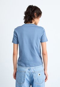 Person med krøllete mørkt hår iført en blå kortermet skjorte og lyseblå denimjeans, sett bakfra mot en hvit bakgrunn.