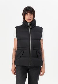 Svart puffer vest med høy krage, frontglidelås, to sidelommer og kviltet design. Brukt over en hvit t-skjorte med korte ermer.