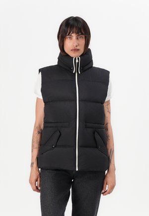 Black puffer vest with high collar, front zipper, two side pockets, and quilted design. Worn over a white t-shirt with short sleeves.