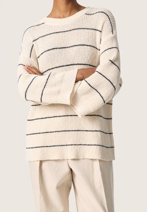 Personne portant un pull en tricot crème avec des rayures horizontales noires et des manches larges, associé à un pantalon ample beige clair.