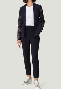 Blazer azul marino con mangas de encaje, camisa blanca y pantalones negros a medida. Combinado con zapatillas plateadas para un look moderno.