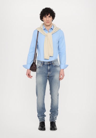 Lichtblauw overhemd met lange mouwen, beige trui drapeert, vervaagde blauwe jeans en zwarte schoenen. Kleine donkerbruine tas in de linkerhand.