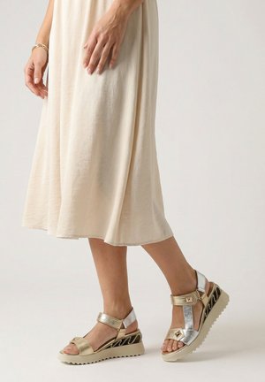 Nogi w srebrno-złotych sandałach na koturnie z paskami na rzep, do tego płynąca kremowa spódnica midi i złota bransoletka.