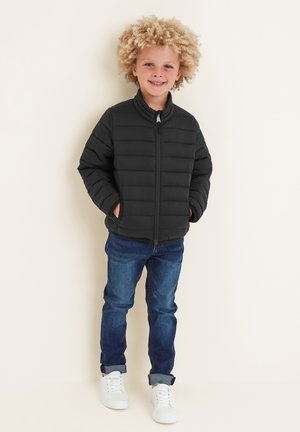 Smiling child with curly blond hair stands against a plain wall, wearing a black puffer jacket, blue jeans, and white sneakers.