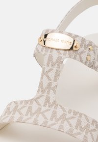 Bride de sandale Michael Kors blanche avec motif MK en relief, plaque logo dorée et rivets sur fond blanc.