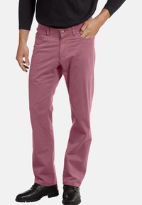 Heren roze katoenen broek met een rechte pijp, vijf zakken en een metalen knoopsluiting. Draagt zwarte schoenen.