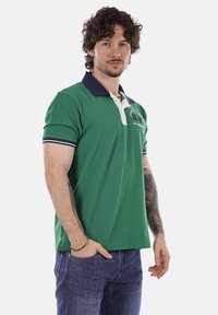 Homme aux cheveux bouclés portant un polo vert avec un col bleu marine et un jean bleu, debout, une main dans la poche, sur un fond blanc.