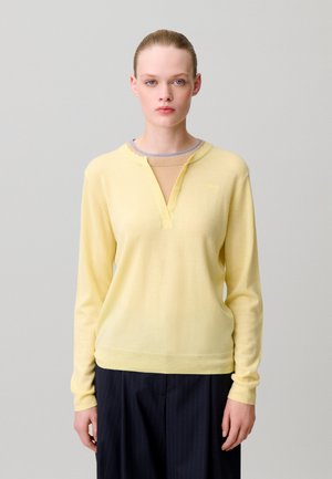 Jeune femme aux cheveux blonds attachés, portant un haut manches longues jaune pâle et un pantalon foncé sur un fond gris uni.