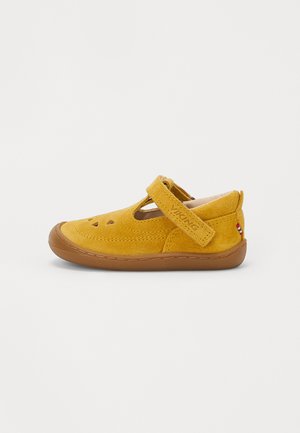 Scarpa di camoscio gialla con punta arrotondata, una fascia in Velcro, dettagli intagliati sui lati e una suola in gomma marrone. Piccolo marchio sulla fascia.