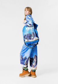 Conjunto azul estampado que incluye una sudadera con capucha y pantalones con diseño gráfico de montañas. Botas marrones y un bolso azul con detalles de cadena completan el conjunto.