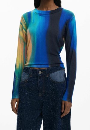 Kobieta ubrana w długi rękaw, wielokolorową bluzkę w odcieniach niebieskiego, zielonego i pomarańczowego oraz wysokiej talii ciemnoniebieskie jeansy w cętki z jasnodżinsowymi kieszeniami.