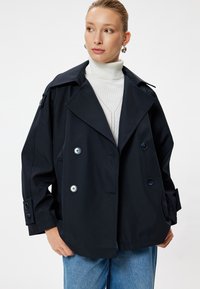 Cappotto trench navy con ampio colletto, design doppiopetto, grandi bottoni e tasche laterali. Indossato sopra un dolcevita crema e denim.