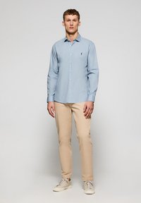 Hellblaues Hemd mit langen Ärmeln und Knopfleiste, das ein dezentes Logo auf der Brust trägt, kombiniert mit beigen Hosen und weißen Sneakern.