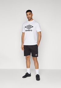 Hvid t-shirt med sort "Umbro" logo, parret med sorte shorts med et lille hvidt logo. Sorte sportsko og hvide sokker.