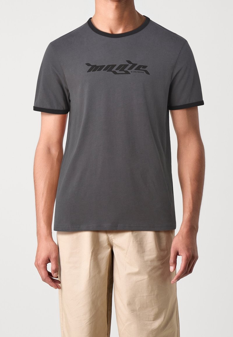 Camiseta gris de cuello redondo con ribete negro en las mangas y el cuello, y con un logo gráfico impreso en el frente. Textura de tela suave.