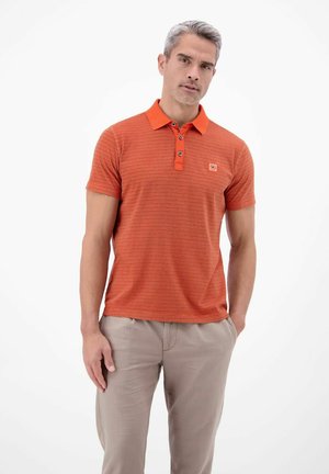 Mann trägt ein orange gestreiftes Poloshirt und beige Hose, steht mit einer Hand in der Tasche vor einem schlichten weißen Hintergrund.