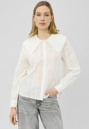 Femme aux cheveux blonds portant une blouse blanche à boutons avec un grand col en dentelle et un jean gris clair, debout avec une main dans la poche.