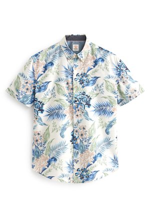 Homme portant une chemise fleurie à manches courtes, un pantalon noir et des chaussures blanches sans lacets, debout devant un fond vert clair uni.
