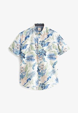 Homme portant une chemise fleurie à manches courtes, un pantalon noir et des chaussures blanches sans lacets, debout devant un fond vert clair uni.