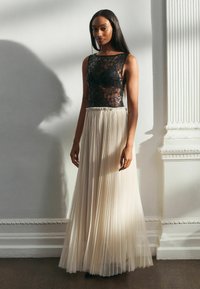 Schwarzer Spitzenoberteil mit semi-transparentem Design, kombiniert mit einem fließenden, plissierten cremfarbenen Rock. Das Outfit hat eine figuroptimierte Taille und eine weiche Textur.