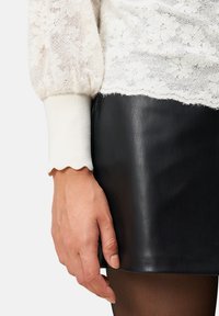 Spitzen-Oberteil mit langen Ärmeln in Off-White mit puffigen Ärmeln und gewellten Bündchen, kombiniert mit einem schwarzen Leder-Minirock. Dunkel durchsichtige Strümpfe sind sichtbar.