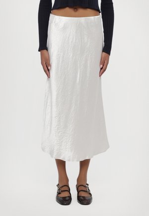 Model draagt een witte gerimpelde midirok, een zwart cropped top met lange mouwen en zwarte platte schoenen met dubbele bandjes, staand tegen een witte achtergrond.