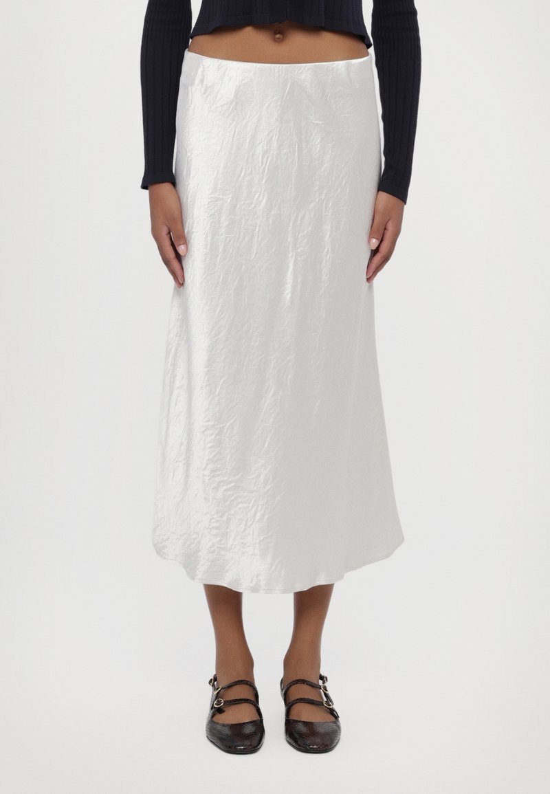Mannequin portant une jupe midi blanche froissée, un haut noir à manches longues raccourci et des chaussures plates noires à double bride, debout devant un fond blanc.