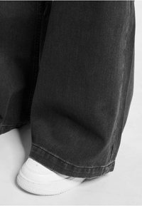 Vide ben bukser i sort denim med en struktureret overflade og afsluttet kant, parret med en hvid sneaker forneden.