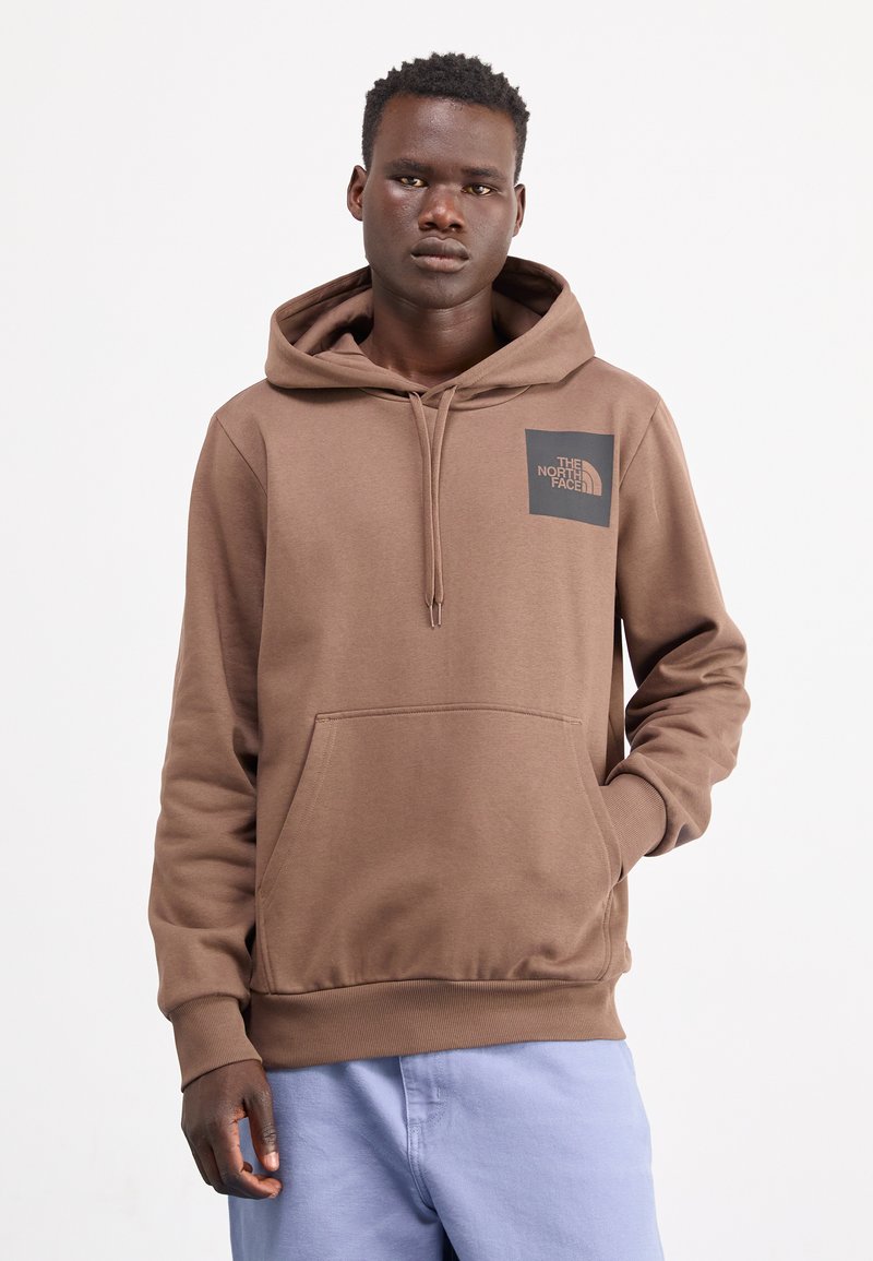 Bruine hoodie met een buidelzak en een zwart vierkant logo op de linkerkant van de borst. Zacht materiaal, ontspannen pasvorm en ribgebreide manchetten.