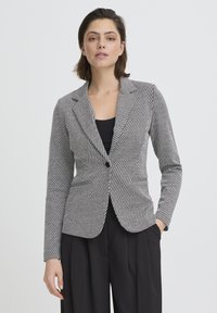 Blazer a righe in tessuto bianco e nero, chiusura con bottone singolo, rever a V, vestibilità sartoriale, maniche lunghe, indossato sopra una maglia nera.