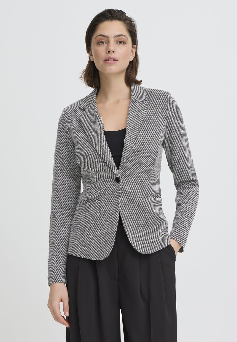 Blazer a righe in tessuto bianco e nero, chiusura con bottone singolo, rever a V, vestibilità sartoriale, maniche lunghe, indossato sopra una maglia nera.