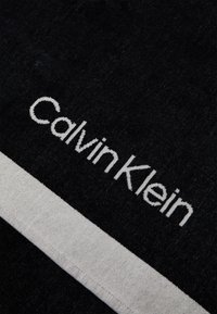 Zwarte stof met een witte horizontale streep waarop "Calvin Klein" in grote letters geborduurd is. Zachte textuur met gelijkmatige stikdetails.