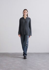 Camisa cinza com riscas, de mangas longas e colarinho com botões, emparelhada com jeans azul escuro e sapatos pretos, de pé contra um fundo neutro.